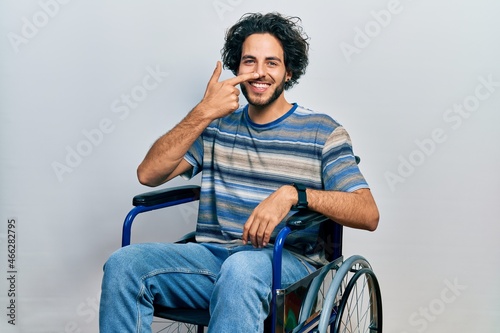 Handsome hispanic man sitting on wheelchair pointing with hand finger to face and nose, smiling cheerful. beauty concept