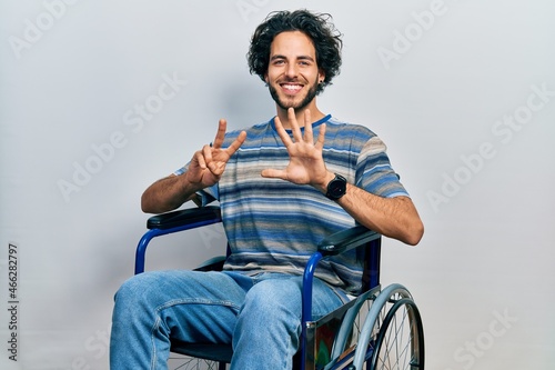 Handsome hispanic man sitting on wheelchair showing and pointing up with fingers number seven while smiling confident and happy.