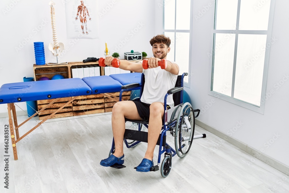 Young arab man patient sitting on wheelchair doing rehab using dumbbells at clinic Young arab man patient sitting on wheelchair doing rehab using dumbbells at clinic