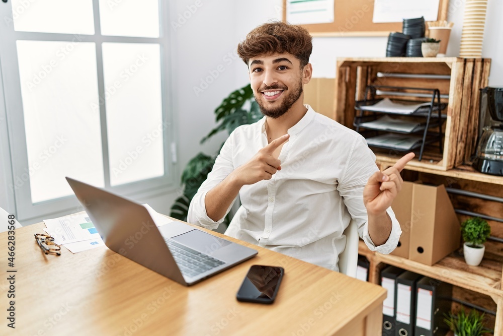 Young arab man working using computer laptop at the office smiling and looking at the camera pointing with two hands and fingers to the side. Young arab man working using computer laptop at the office smiling and looking at the camera pointing with two hands and fingers to the side.