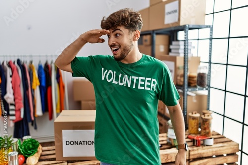 Young arab man wearing volunteer t shirt at donations stand very happy and smiling looking far away with hand over head. searching concept.