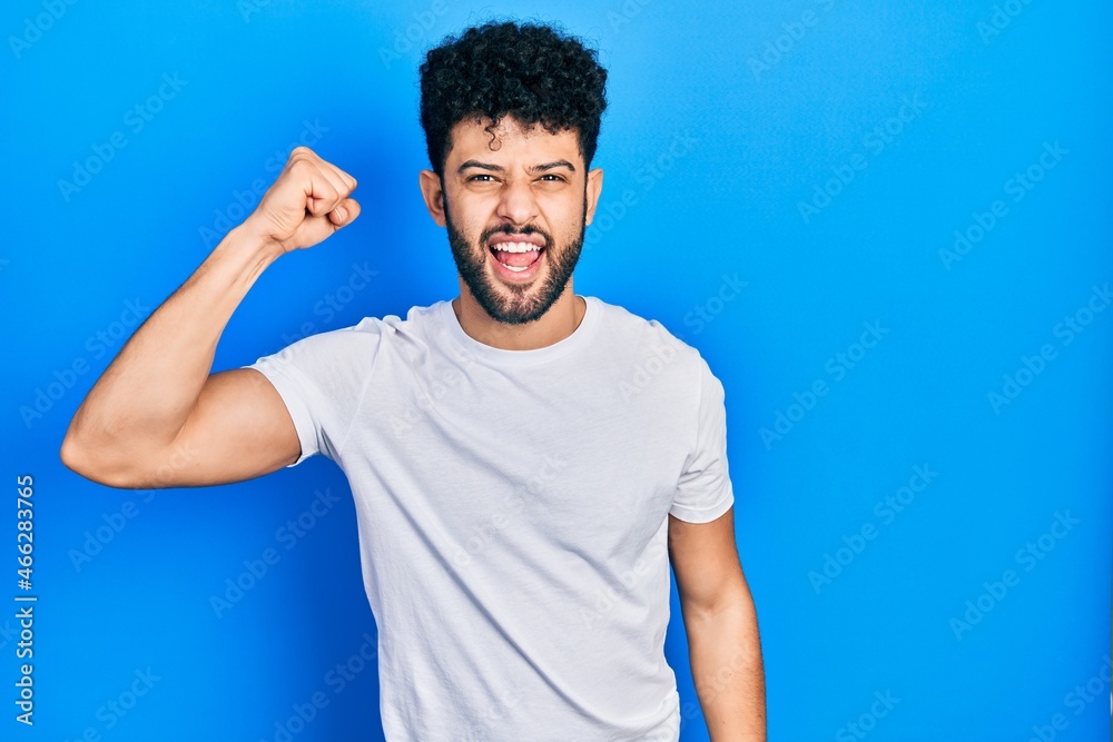 Young arab man with beard wearing casual white t shirt angry and mad raising fist frustrated and furious while shouting with anger. rage and aggressive concept. Young arab man with beard wearing casual white t shirt angry and mad raising fist frustrated and furious while shouting with anger. rage and aggressive concept.