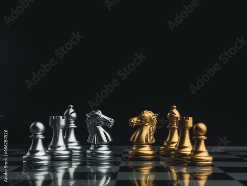 Confrontation of a golden and a silver team of chess pieces, knight, bishop, rook, and pawn on a chessboard on dark background. Leadership, partnership, team, competitor and business strategy concept.