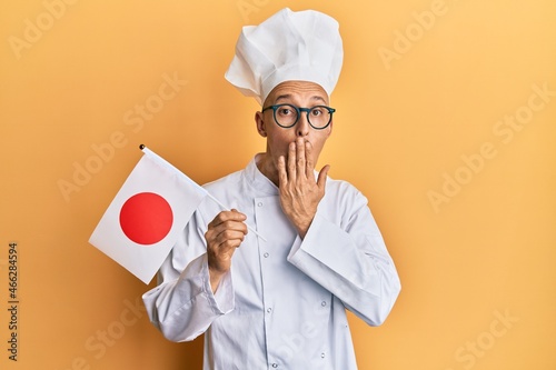 Bald man with beard wearing professional cook uniform holding japan flag covering mouth with hand, shocked and afraid for mistake. surprised expression photo