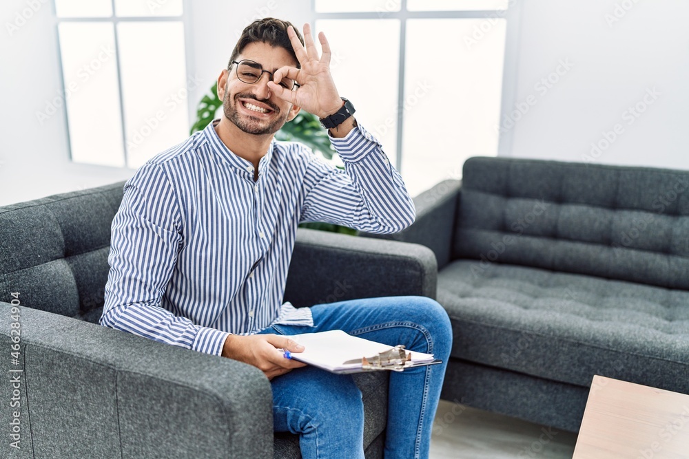 Young psychologist man at consultation office doing ok gesture with hand smiling, eye looking through fingers with happy face. Young psychologist man at consultation office doing ok gesture with hand smiling, eye looking through fingers with happy face.