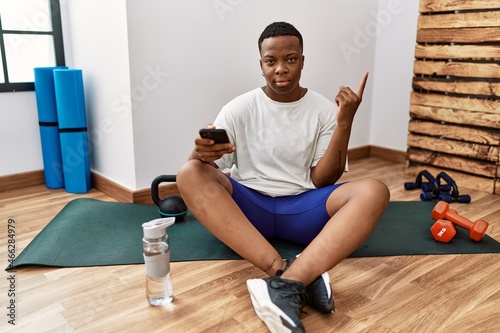 Young african man sitting on training mat at the gym using smartphone pointing with hand finger to the side showing advertisement  serious and calm face