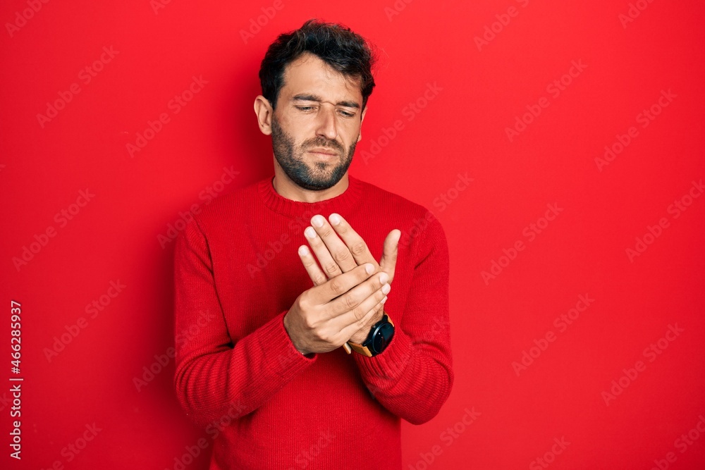 Handsome man with beard wearing casual red sweater suffering pain on hands and fingers, arthritis inflammation