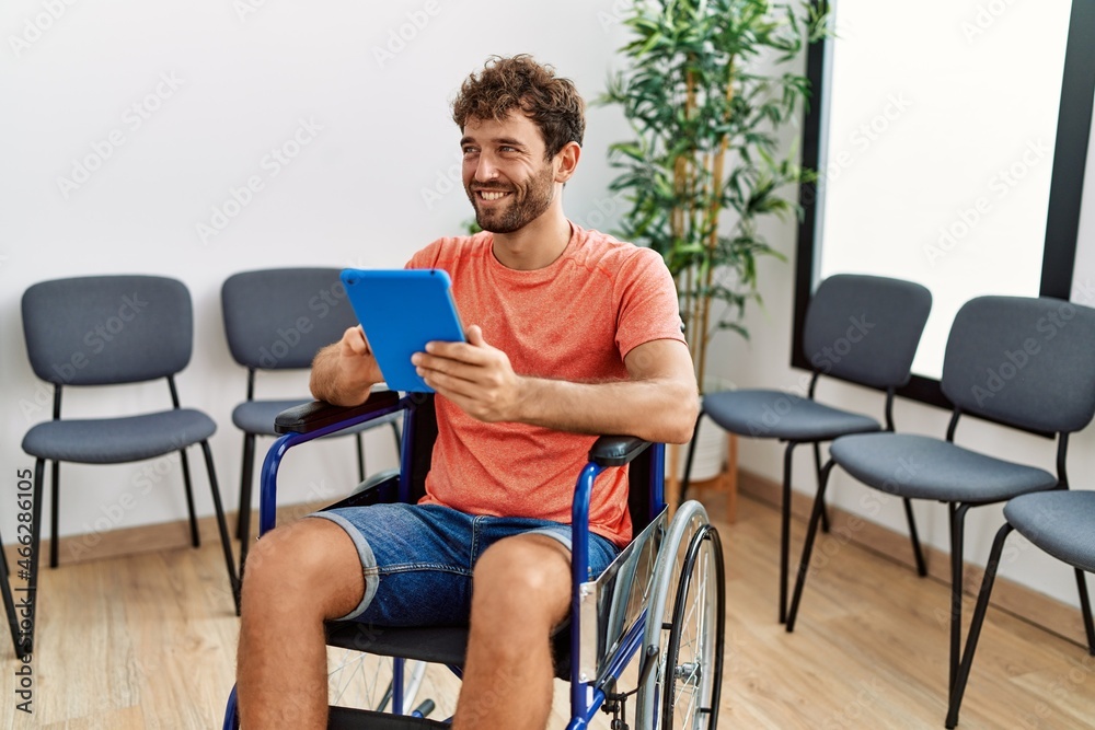 Young hispanic man using touchpad sitting on wheelchair at clinic waiting room. Young hispanic man using touchpad sitting on wheelchair at clinic waiting room.