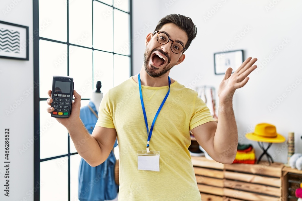 Young man with beard working at retail boutique holding dataphone celebrating achievement with happy smile and winner expression with raised hand Young man with beard working at retail boutique holding dataphone celebrating achievement with happy smile and winner expression with raised hand