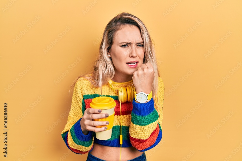 Beautiful young blonde woman drinking cup of coffee wearing headphones angry and mad raising fist frustrated and furious while shouting with anger. rage and aggressive concept. Beautiful young blonde woman drinking cup of coffee wearing headphones angry and mad raising fist frustrated and furious while shouting with anger. rage and aggressive concept.