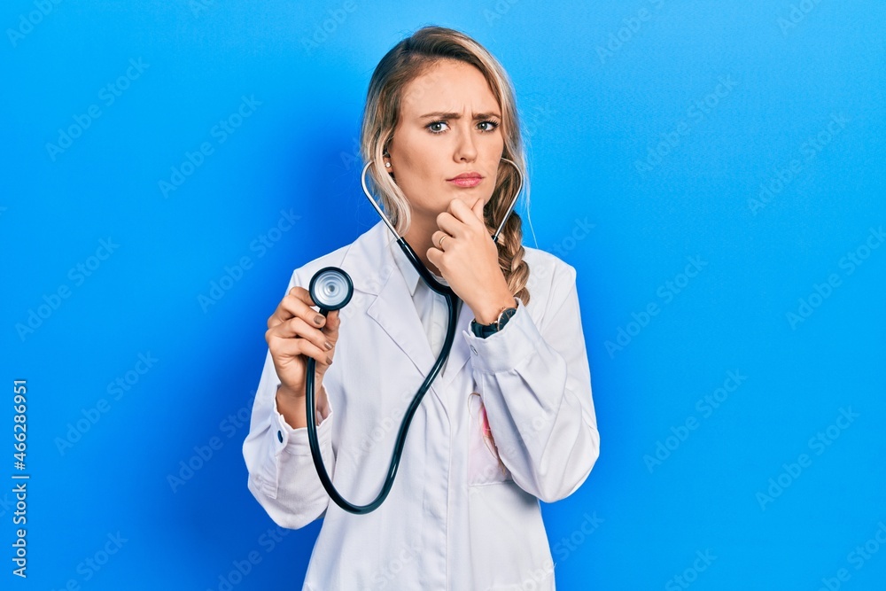 Beautiful young blonde doctor woman holding stethoscope thinking worried about a question, concerned and nervous with hand on chin Beautiful young blonde doctor woman holding stethoscope thinking worried about a question, concerned and nervous with hand on chin