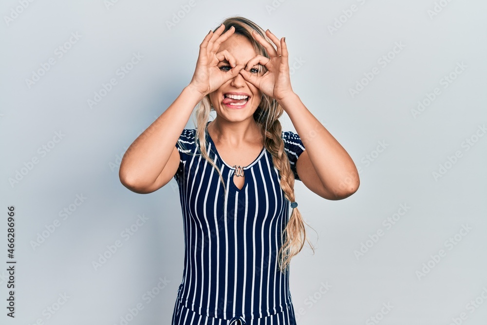 Beautiful young blonde woman wearing casual striped dress doing ok gesture like binoculars sticking tongue out, eyes looking through fingers. crazy expression. Beautiful young blonde woman wearing casual striped dress doing ok gesture like binoculars sticking tongue out, eyes looking through fingers. crazy expression.