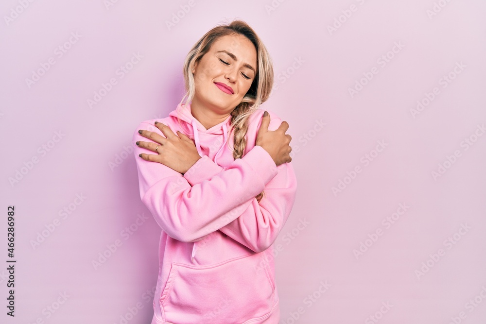Beautiful young blonde woman wearing pink sweatshirt hugging oneself happy and positive, smiling confident. self love and self care Beautiful young blonde woman wearing pink sweatshirt hugging oneself happy and positive, smiling confident. self love and self care