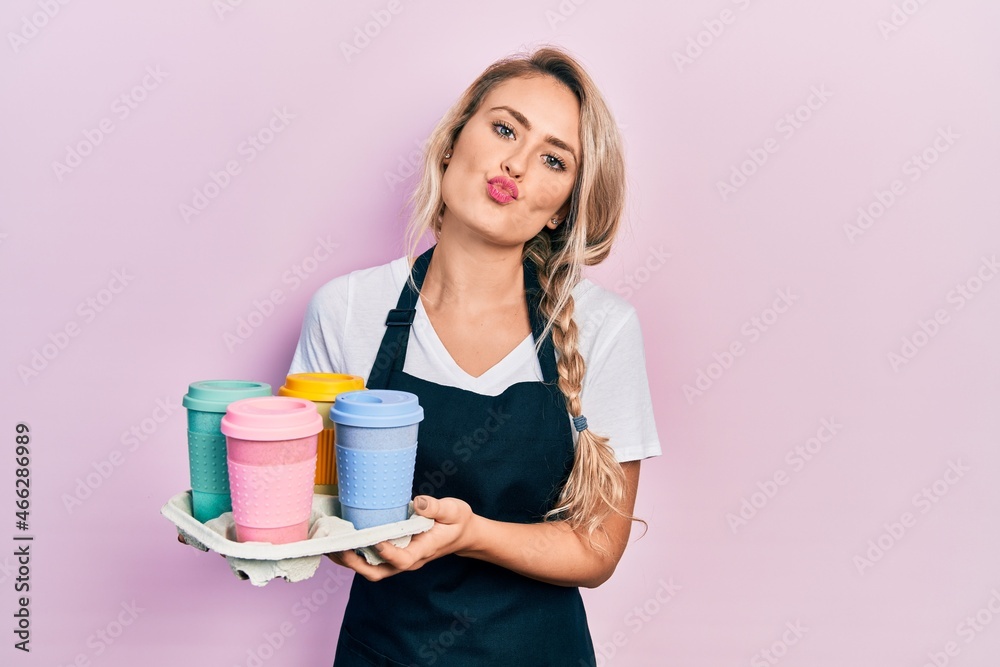 Beautiful young blonde woman wearing waitress apron holding take away cup of coffees looking at the camera blowing a kiss being lovely and sexy. love expression. Beautiful young blonde woman wearing waitress apron holding take away cup of coffees looking at the camera blowing a kiss being lovely and sexy. love expression.