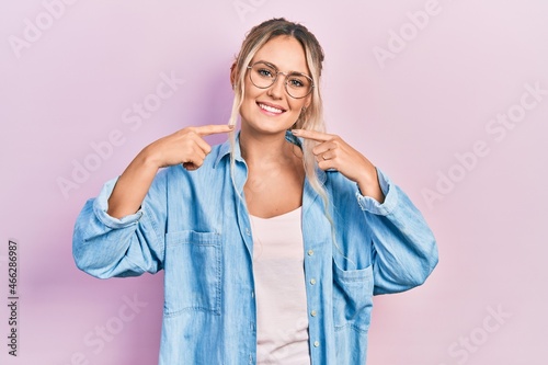 Beautiful young blonde woman wearing casual clothes and glasses smiling cheerful showing and pointing with fingers teeth and mouth. dental health concept. © Krakenimages.com