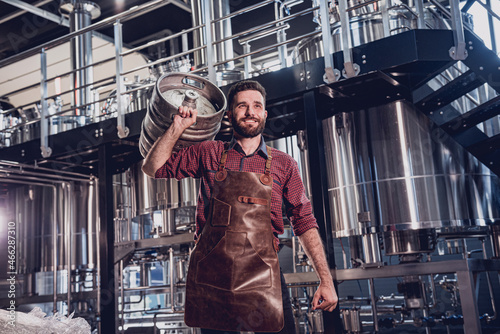 Young male brewer in leather apron holds barrel with craft beer at modern brewery factory