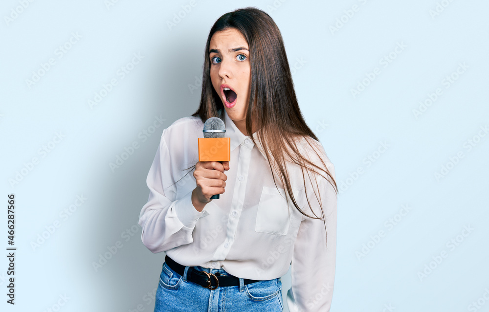 Young brunette teenager holding reporter microphone scared and amazed with open mouth for surprise, disbelief face Young brunette teenager holding reporter microphone scared and amazed with open mouth for surprise, disbelief face