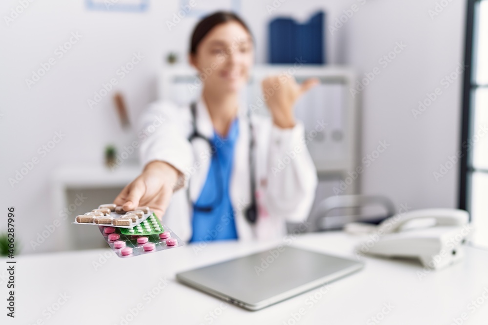 Young hispanic doctor woman wearing doctor uniform holding prescription pills at clinic pointing thumb up to the side smiling happy with open mouth Young hispanic doctor woman wearing doctor uniform holding prescription pills at clinic pointing thumb up to the side smiling happy with open mouth