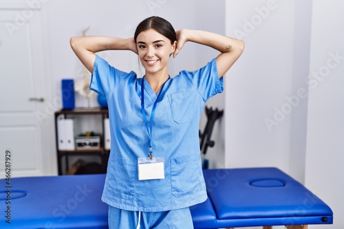 Young hispanic woman wearing physiotherapist uniform standing at clinic relaxing and stretching  arms and hands behind head and neck smiling happy