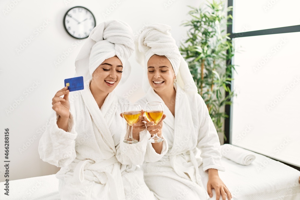 Woman couple holding credit card and toasting with champagne sitting on massage board at beauty center