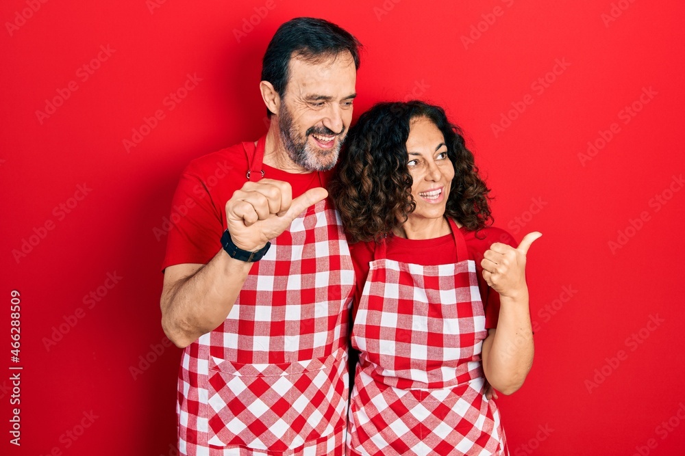 Middle age couple of hispanic woman and man wearing cook apron smiling with happy face looking and pointing to the side with thumb up. Middle age couple of hispanic woman and man wearing cook apron smiling with happy face looking and pointing to the side with thumb up.