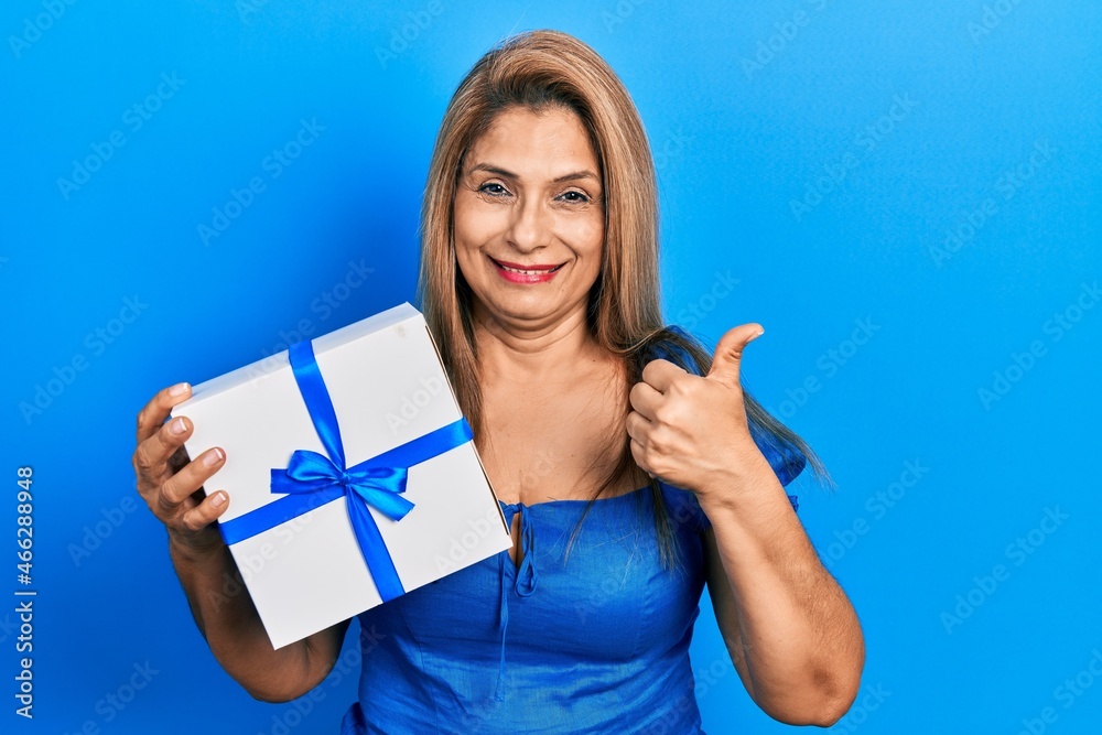Middle age hispanic woman holding gift smiling happy and positive, thumb up doing excellent and approval sign Middle age hispanic woman holding gift smiling happy and positive, thumb up doing excellent and approval sign