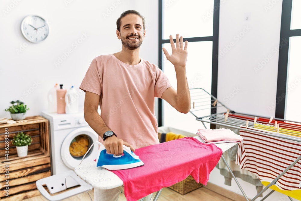 Young hispanic man ironing clothes at home waiving saying hello happy and smiling, friendly welcome gesture