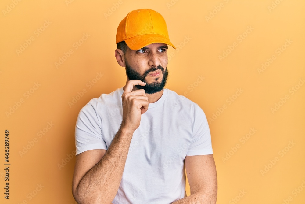 Young man with beard wearing yellow cap thinking concentrated about doubt with finger on chin and looking up wondering Young man with beard wearing yellow cap thinking concentrated about doubt with finger on chin and looking up wondering