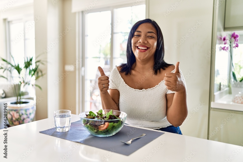 Young hispanic woman eating healthy salad at home success sign doing positive gesture with hand, thumbs up smiling and happy. cheerful expression and winner gesture.