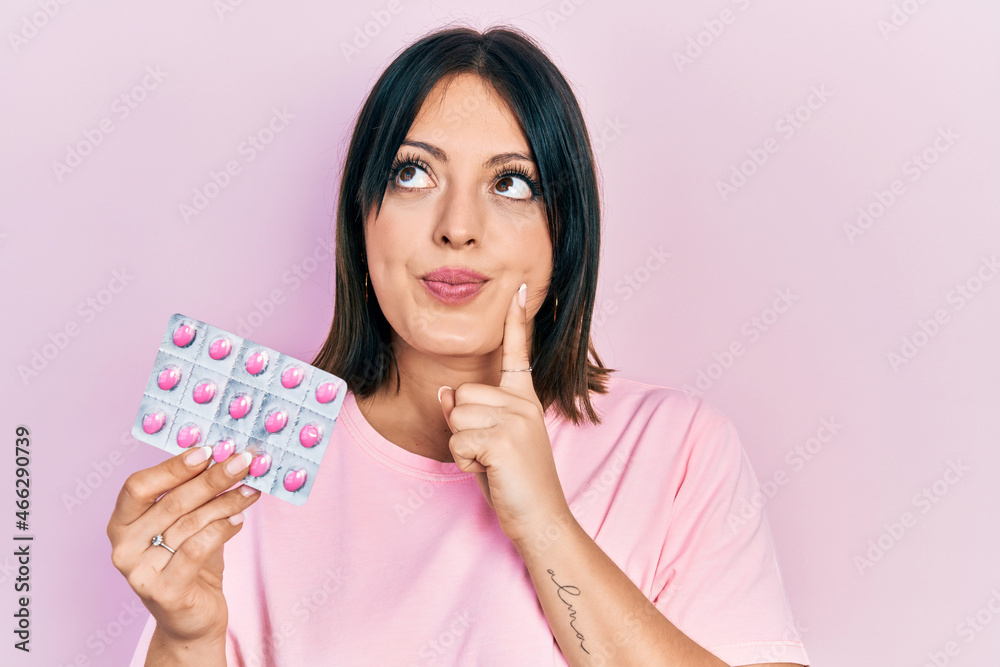 Young hispanic woman holding pills serious face thinking about question with hand on chin, thoughtful about confusing idea Young hispanic woman holding pills serious face thinking about question with hand on chin, thoughtful about confusing idea