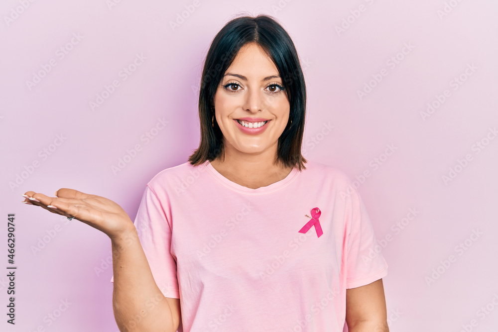 Young hispanic woman wearing pink cancer ribbon on t shirt smiling cheerful presenting and pointing with palm of hand looking at the camera. Young hispanic woman wearing pink cancer ribbon on t shirt smiling cheerful presenting and pointing with palm of hand looking at the camera.