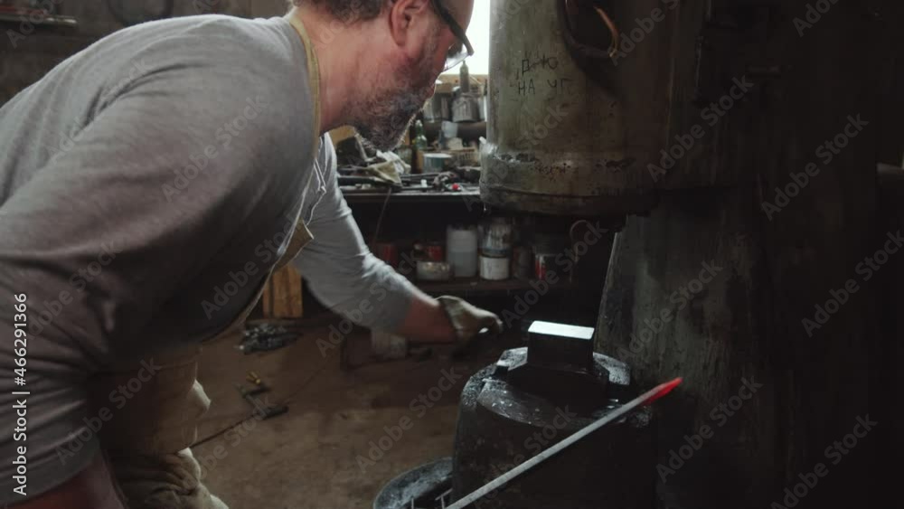 Blacksmith in protective gloves forging heated iron rod with power hammer machine in workshop Blacksmith in protective gloves forging heated iron rod with power hammer machine in workshop