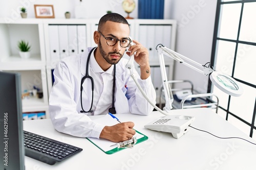 Young hispanic man wearing doctor uniform speaking on the phone at clinic