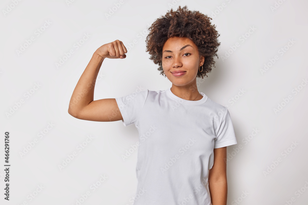 Confident woman feels strength and power raises arm flexes biceps and looks proud of her own achievements has strong muscles wears casual t shirt isolated over white background brags with fit body Confident woman feels strength and power raises arm flexes biceps and looks proud of her own achievements has strong muscles wears casual t shirt isolated over white background brags with fit body