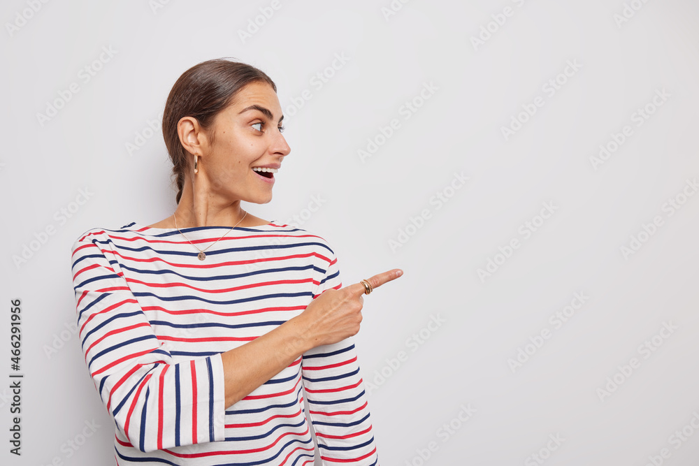 Beautiful surprised woman points index finger away looks at advertisement shows empty space for your promotonal content wears casual striped jumper isolated over white background. Look at this Beautiful surprised woman points index finger away looks at advertisement shows empty space for your promotonal content wears casual striped jumper isolated over white background. Look at this