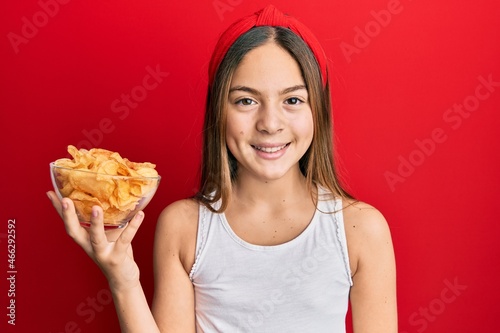 Beautiful brunette little girl holding potato chips looking positive and happy standing and smiling with a confident smile showing teeth