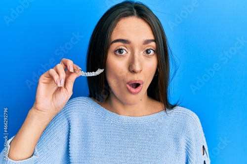 Young hispanic girl holding invisible aligner orthodontic scared and amazed with open mouth for surprise, disbelief face