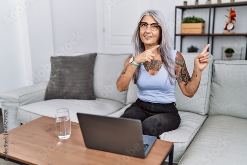 Middle age grey-haired woman using laptop at home smiling and looking at the camera pointing with two hands and fingers to the side.