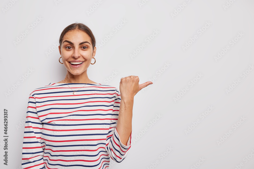 Horizontal shot of positive young woman points thumb away shows blank space demonstrates advertisement or promotion wears casual striped jumper smiles positively isolated over white background Horizontal shot of positive young woman points thumb away shows blank space demonstrates advertisement or promotion wears casual striped jumper smiles positively isolated over white background
