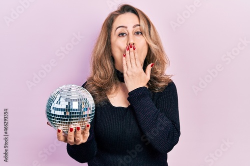 Middle age caucasian woman holding shiny disco ball covering mouth with hand, shocked and afraid for mistake. surprised expression photo