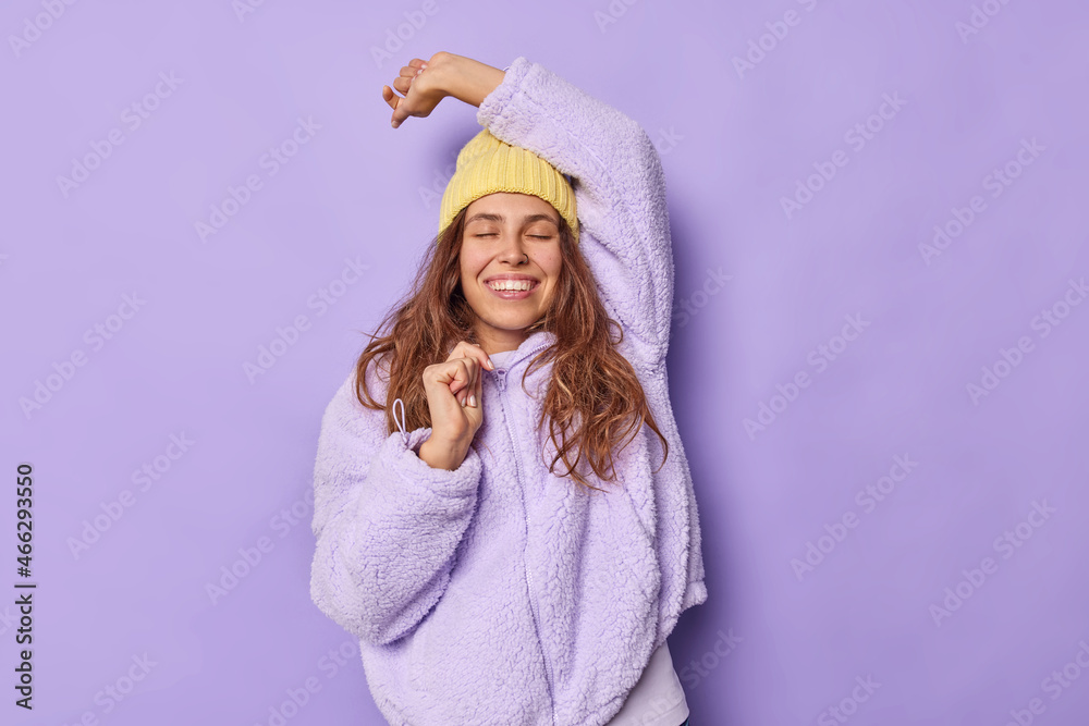 Pleased young woman keeps eye closed dances carefree keeps arm raised up wears yellow hat and warm jacket enjoys life isolated over purple background. People joy and pleasant feelings concept Pleased young woman keeps eye closed dances carefree keeps arm raised up wears yellow hat and warm jacket enjoys life isolated over purple background. People joy and pleasant feelings concept