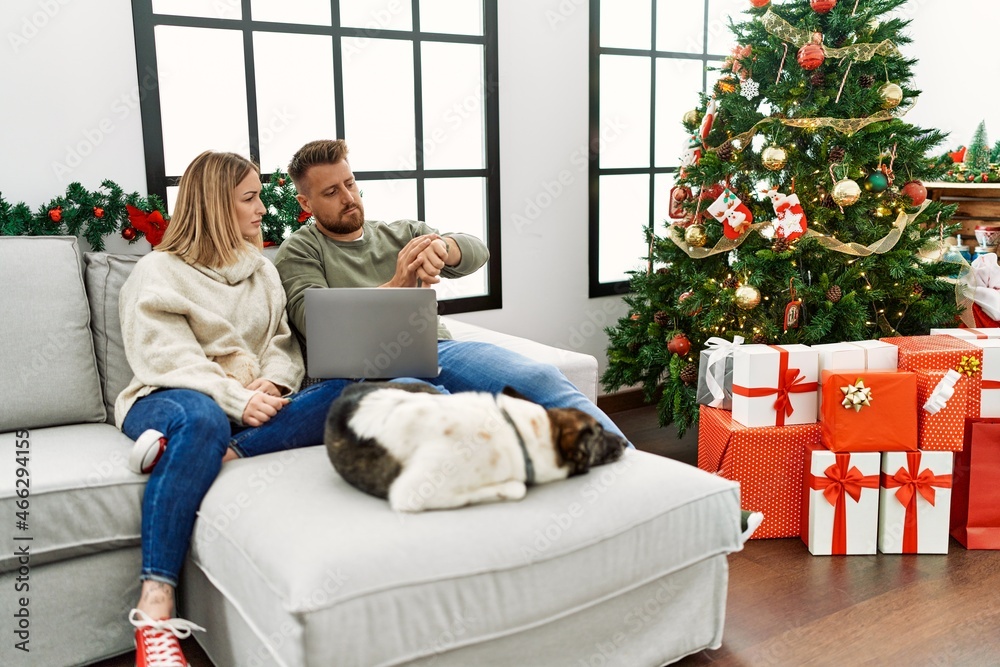 Young couple using laptop sitting by christmas tree checking the time on wrist watch, relaxed and confident