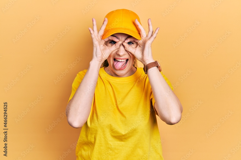 Young hispanic woman wearing delivery uniform and cap doing ok gesture like binoculars sticking tongue out, eyes looking through fingers. crazy expression.
