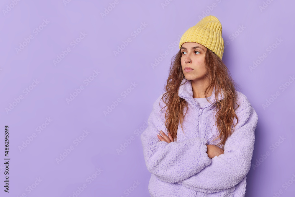 Thoughtful good looking female teenager has long hair wears yellow hat and fur coat keeps arms folded concentrated away poses against purple background with copy space for your advertisement Thoughtful good looking female teenager has long hair wears yellow hat and fur coat keeps arms folded concentrated away poses against purple background with copy space for your advertisement