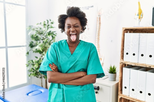 African young physiotherapist woman working at pain recovery clinic sticking tongue out happy with funny expression.