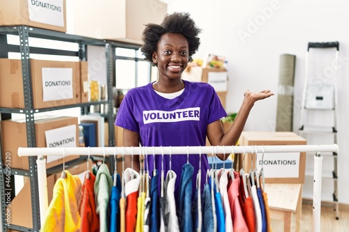 African young woman wearing volunteer t shirt at donations stand smiling cheerful presenting and pointing with palm of hand looking at the camera.
