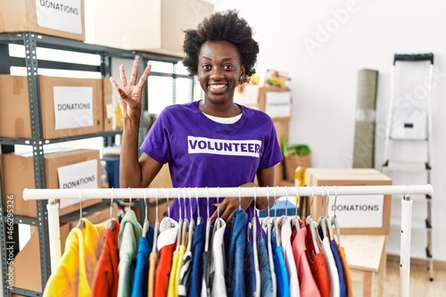 African young woman wearing volunteer t shirt at donations stand showing and pointing up with fingers number four while smiling confident and happy.