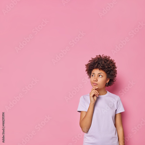 Vertical shot of good looking woman with curly hair holds chin thinks deeply about somehing considers something wears casual clothes poses against pink background blank copy space for advert