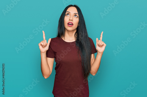 Young hispanic girl wearing casual t shirt amazed and surprised looking up and pointing with fingers and raised arms.