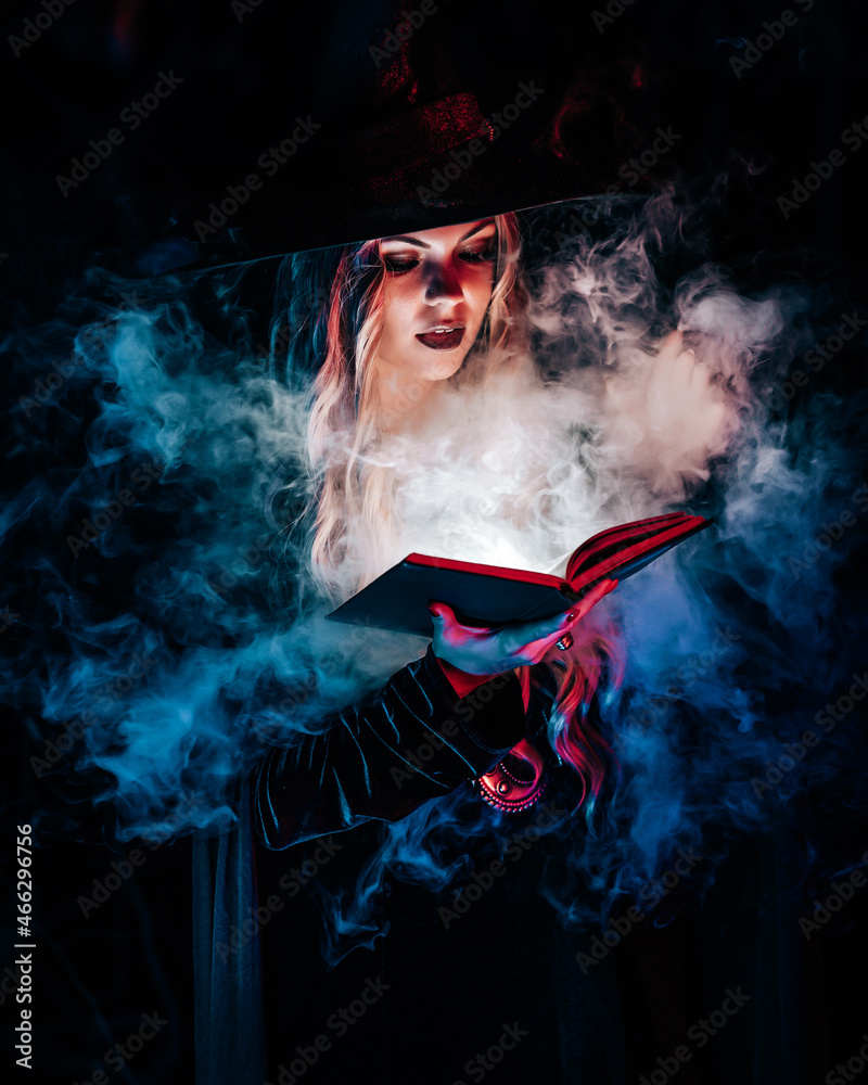 Beautiful witch reading magic book on dark background in forest. Halloween, blonde wizard costume concept. Pretty woman in blue smoke. 